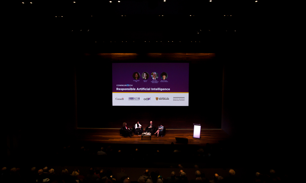 Far shot of the entire stage with panelists centered and a presentation playing