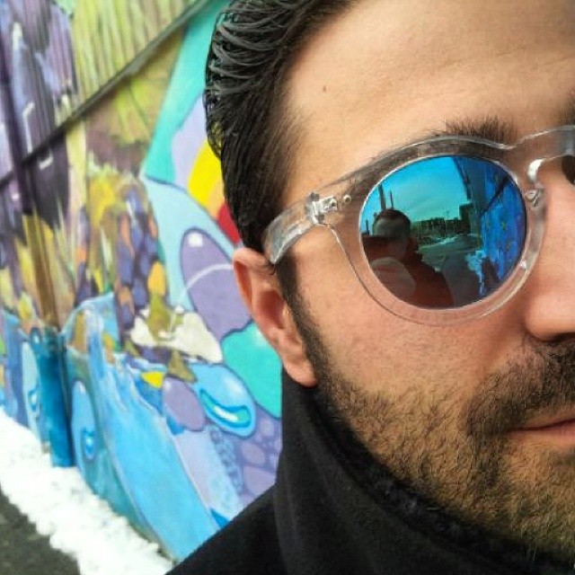 A close up shot of half a persons face wearing blue sunglasses. There is colourful street art behind.