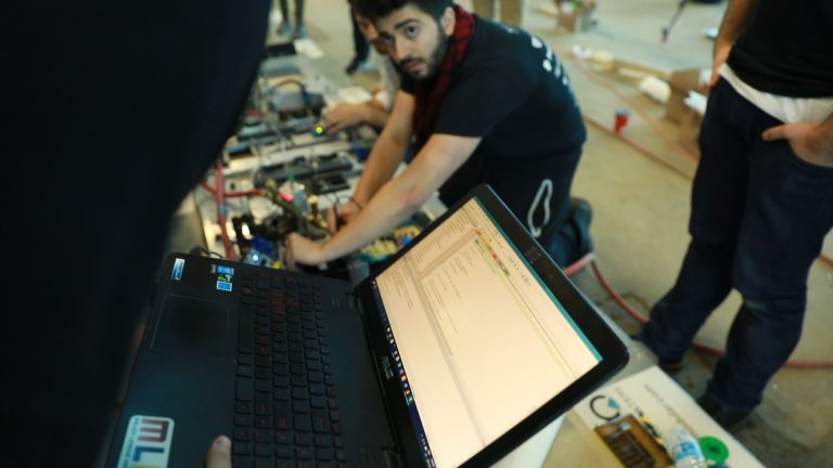 A close up of a computer screen with people working on electronics in the background.